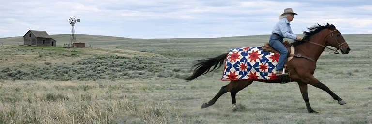 Cowboy riding horse with a handmade quilt across a prairie landscape